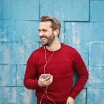 A joyful man in a red sweater smiles while listening to music on his smartphone against a blue brick wall.